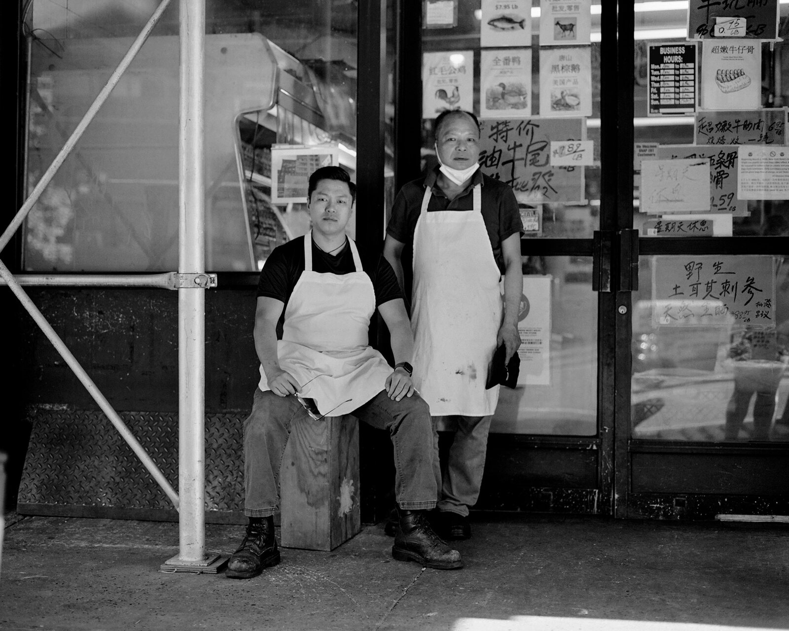 Jefferson Li seated and his father Peter standing outside the shop, hand-written Chinese price signs filling the window behind them